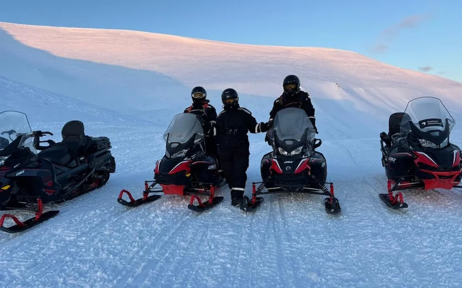 Four parked snowmobiles and people in snowsuits on an open white snowfield