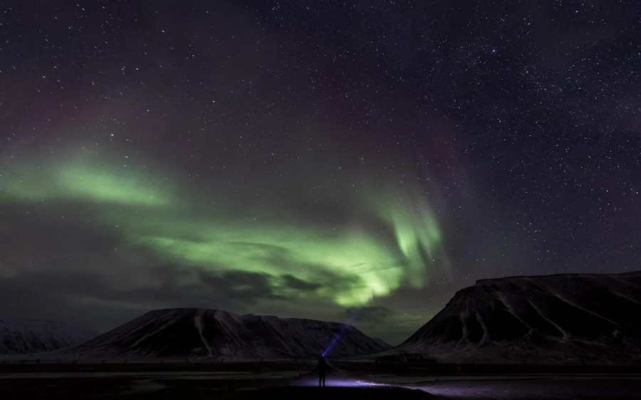 Green northern lights curling across a starry sky above dark mountains