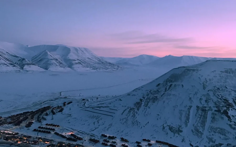 A pink and violet Arctic sky over snowy ridges with the lights of the town below
