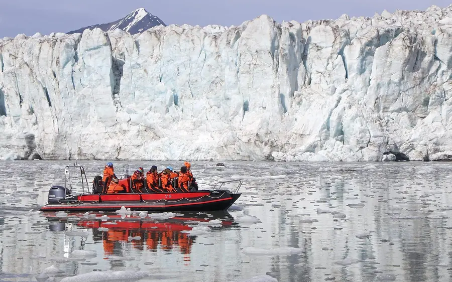A red inflatable boat with passengers in survival suits in front of a tall white glacier wall